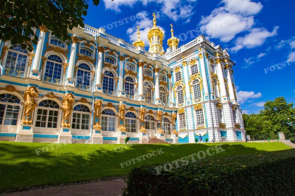 На фотографии изображена часть Екатерининского дворца в Царском Селе, расположенного в городе Пушкин, Санкт-Петербург. Это барочное здание с ярко выраженными золотыми украшениями на фасаде и изящными статуями, которое является примером роскошной императорской архитектуры 18-го века. На переднем плане виден ухоженный зелёный газон, а синее небо с белыми облаками идеально контрастирует с золотистыми и голубыми оттенками дворца. Фотография подходит для исторических материалов, путеводителей, культурно-образовательных изданий и промоционных брошюр, посвящённых русскому барокко и туризму в России.